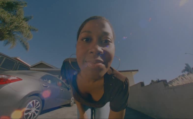 A young black woman bends over and looks directly into the camera and there is a silver automobile behind her