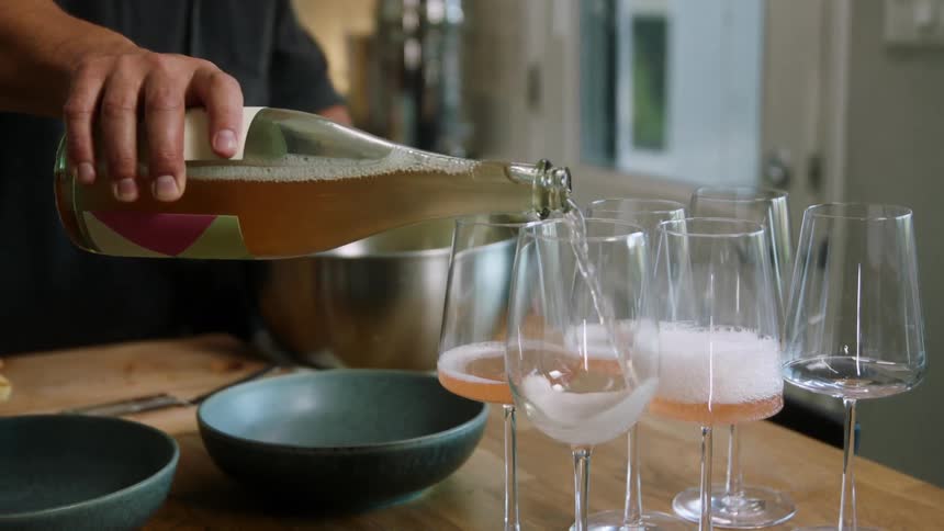 A man pours bubbly wine into several glasses on a wooden kitchen counter
