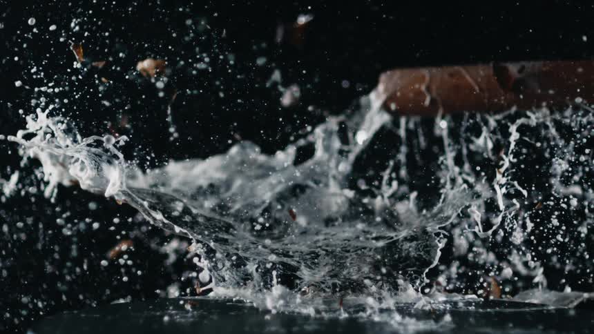 A glass bottle sits on a table as a baseball bat smashes it and liquid splashes and flies
