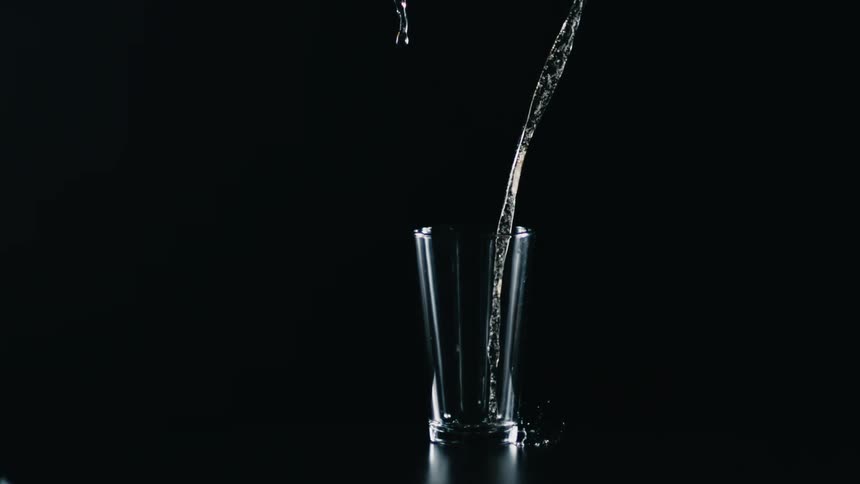 A clear drinking glass is on a table in the darkness as water falls from above and into the glass