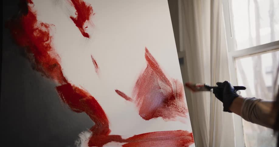 An artist uses a paintbrush to paint on a canvas while standing indoors during the day
