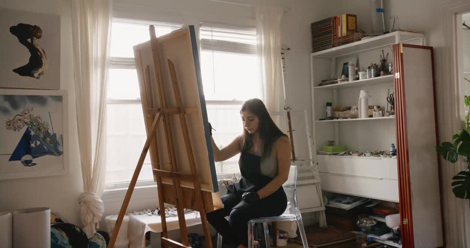 An artist paints on a large canvas on an easel while sitting in a chair indoors during the day