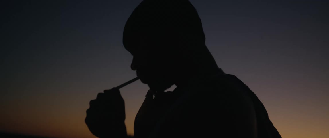 Man with beanie and lighting up a cigarette is silhouetted against the sky