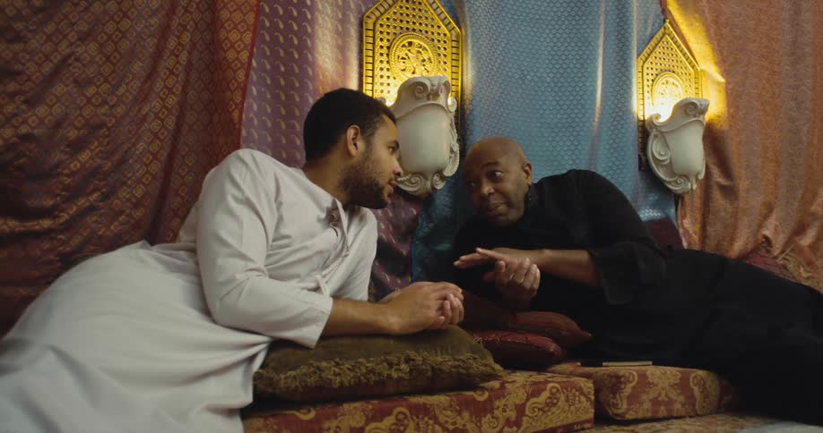 Two men lie on pillows stacked in room and one man talks to the other as he moves his hands a lot