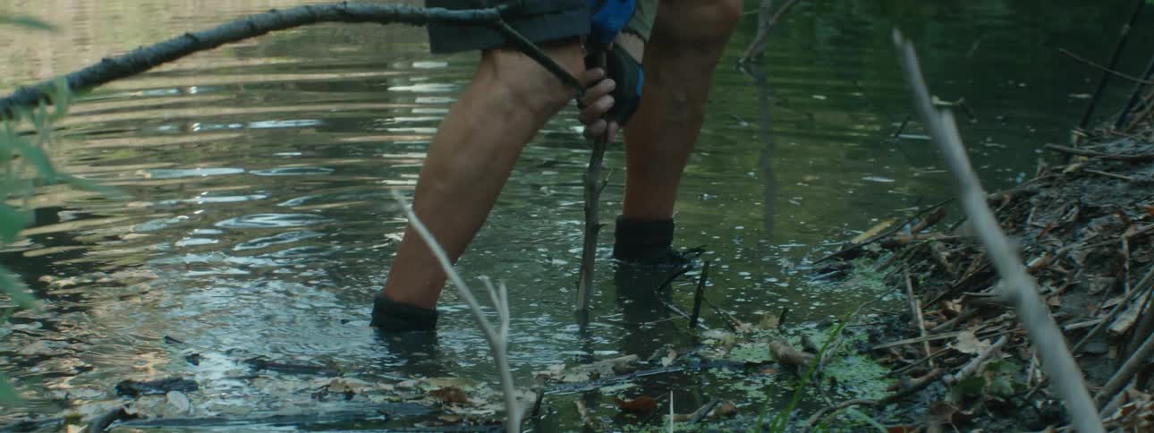 An elderly man stands in a river and pushes a stick in to the bottom of the river