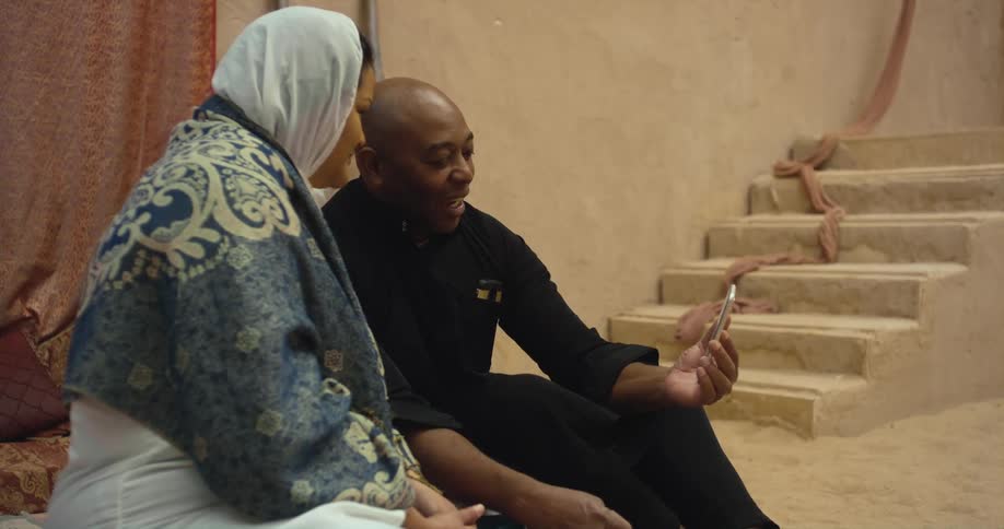 Man wearing black shirt and pants sits next to woman with white headscarf using smartphone and making video call