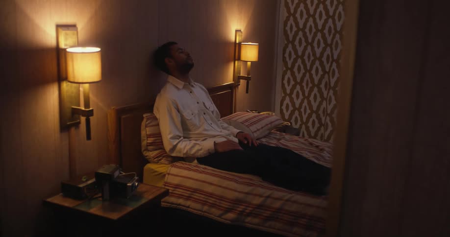 A depressed man rests his head against the wall while sitting in bed