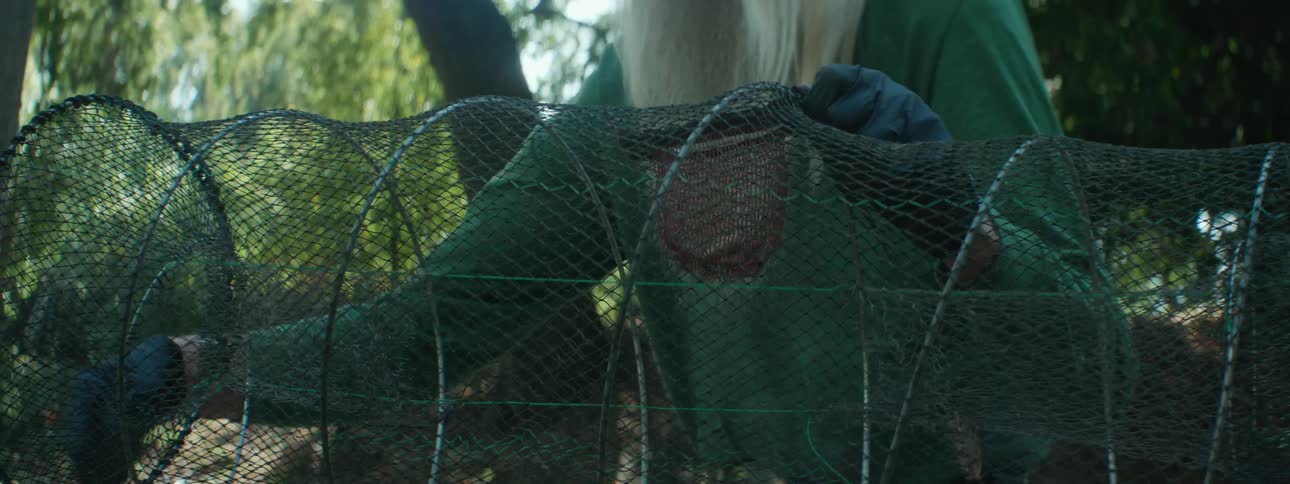 A man holding up a fishing cage uses his hand to explain how it works