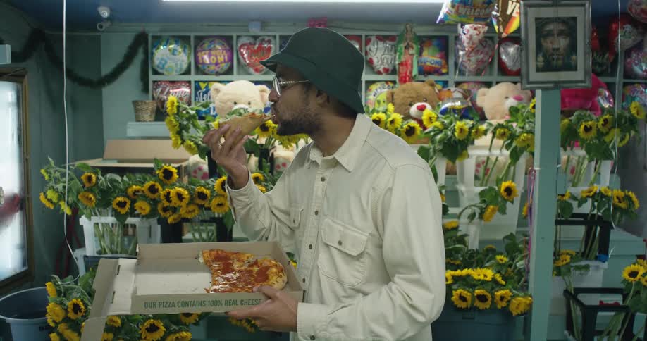 Man stands looking around flower shop while he eats a piece of pizza from the box he is holding