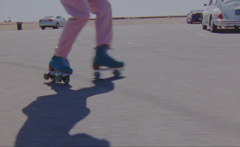 A man wearing pink pants is dancing on blue roller skates in a parking lot