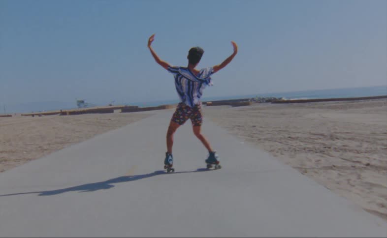 A man wearing shorts and a striped shirt is dancing on roller skates on a road at the beach