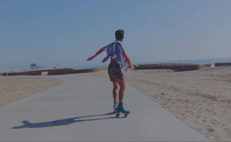 A man wearing shorts and a striped shirt is dancing on roller skates on a road at the beach