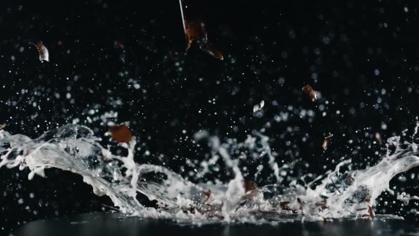 A beer bottle sits on a table and a blade hits it and smashes it and the beer flies everywhere