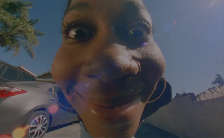 A woman bends forward to put her smiling face close to the camera before backing away and waving while standing in the driveway of a house during the day