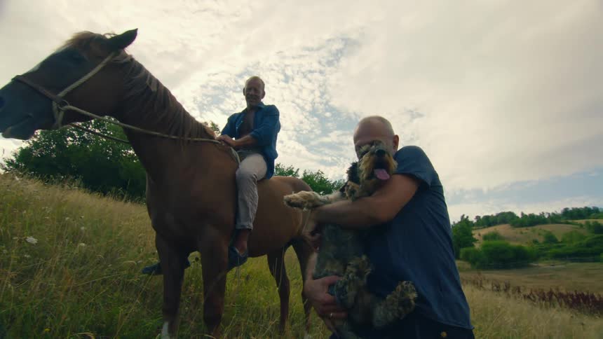 A man sitting atop a horse and a man holding a dog in his arms look into the camera while on a hill in the countryside during the day