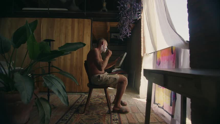 Shirtless man with shorts sits in chair shaving and holding mirror