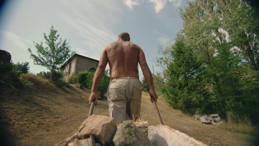 A shirtless man pulls a wagon filled with stones up a hill towards a building during the day