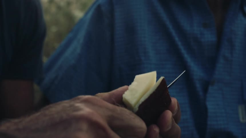 Man cuts a piece of melon next to man wearing blue shirt outdoors