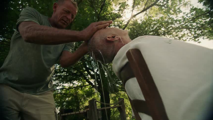 A man sitting leaning back in a chair in the garden while another man scoops water with his hands and washes his hair
