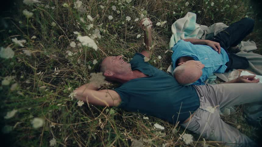 A pair of men laugh while resting together in a field of tall grass during the day