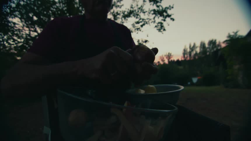 A man peels a bowl of potatoes while sitting outdoors in the evening