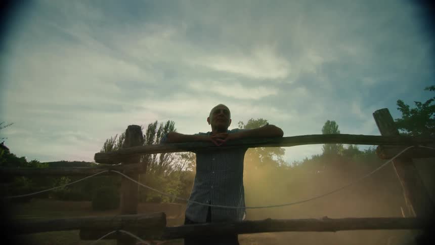 Man stands leaning against top of wooden corral backlit by sunlight
