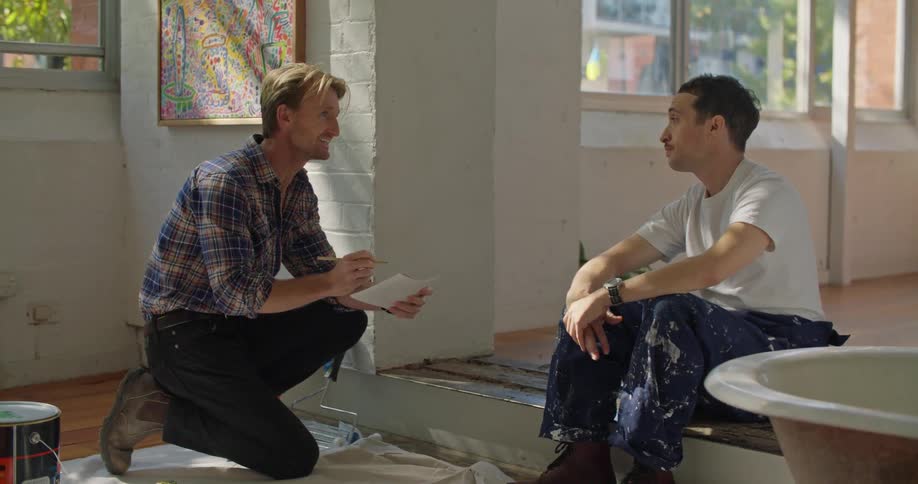 A man writes on a piece of paper while talking to another man sitting nearby before the two men laugh and fist bump