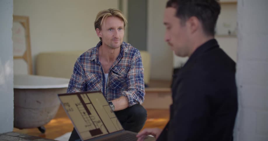 A man sitting on the floor while viewing a floor plan on a laptop talks with a kneeling man taking notes on a sheet of paper before the two shake hands and rise to their feet