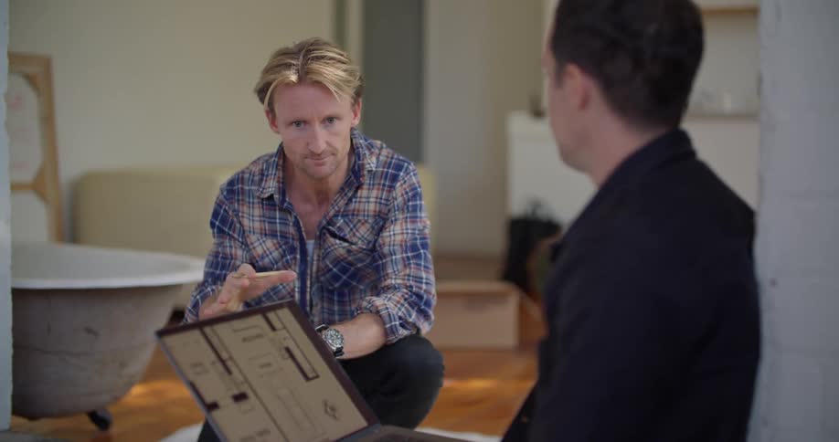 A kneeling man gestures with his hands as he talks with a man sitting nearby with a laptop