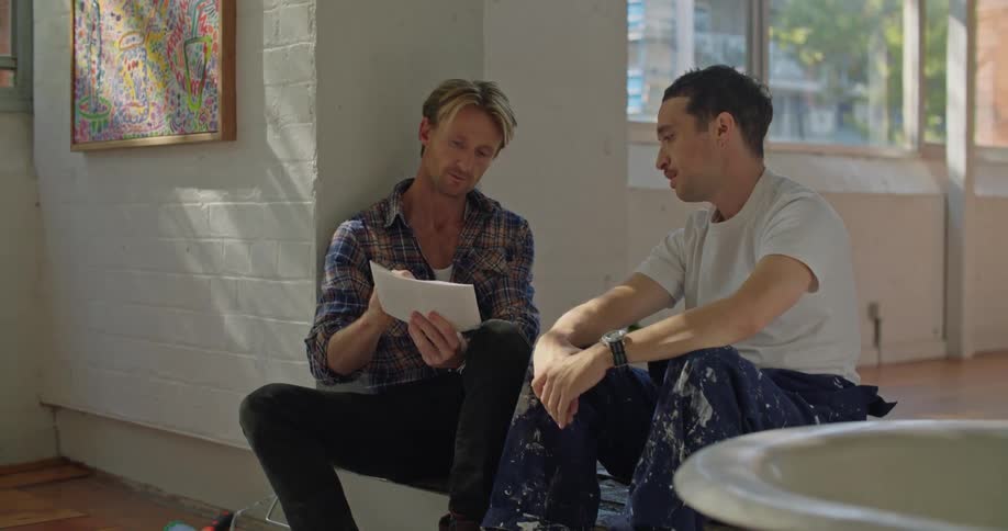 A man shows a sheet of paper to a man sitting beside him and gestures around the room as the two men speak
