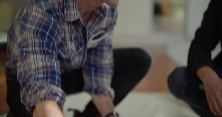 A kneeling man uses a measuring tape to measure a section of flooring while talking to another man kneeling beside him indoors during the day