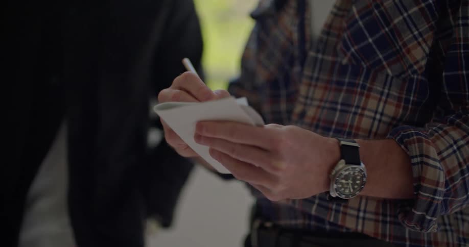 A man writes on a folded piece of paper while another person watches from his side