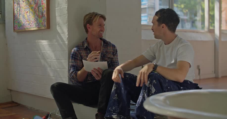 Two men are sitting inside on a step and one man is holding a paper and the other man is playing with a pencil and he gives it back to the other man