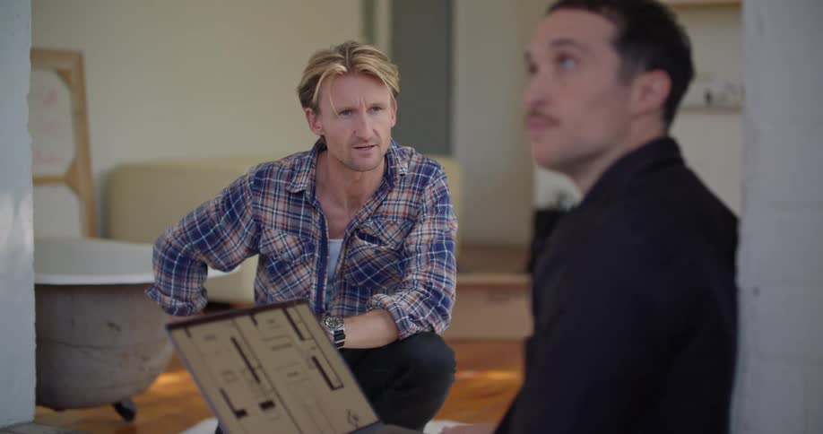 A man writing on a folded sheet of paper gestures while speaking to another man checking a floor plan on a laptop