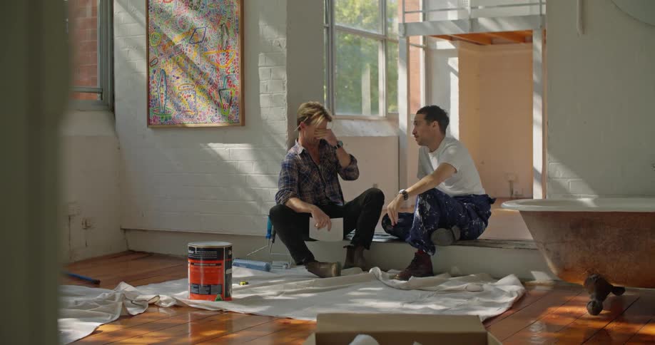 A pair of men speak with one another while sitting together on the floor of an empty house