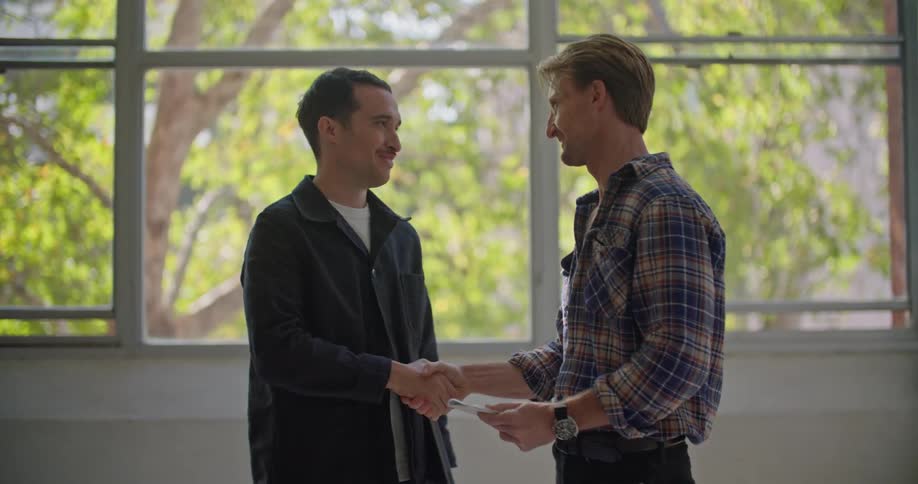Two men are standing inside near windows talking as one man writes on paper and they shake hands and leave