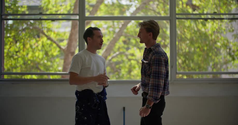 A pair of men speak with one another while gesturing at their surroundings before one man leads the other out of frame