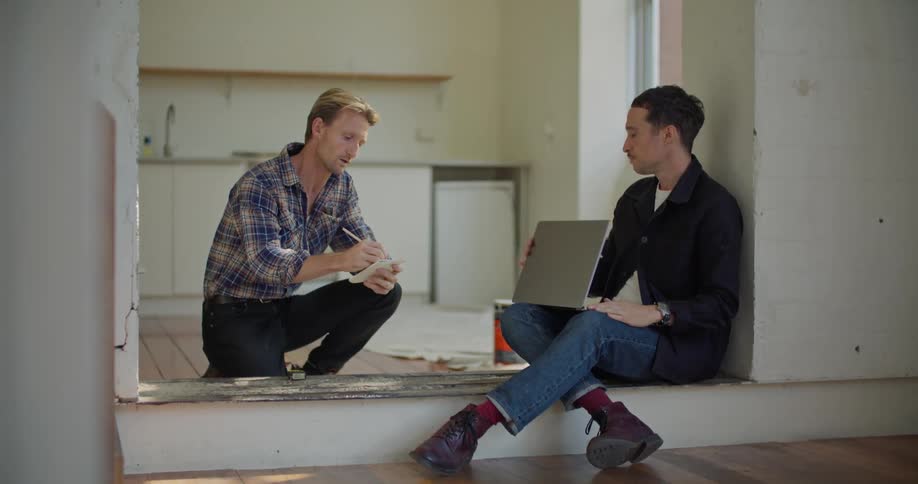 A man is sitting on a step with a laptop computer and another man is on one knee talking to him and writing on a notepad