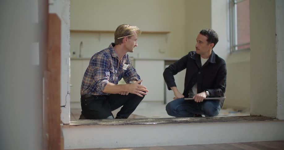 A kneeling man uses a measuring tape to measure a section of floor while speaking with another man kneeling beside him in a house during the day