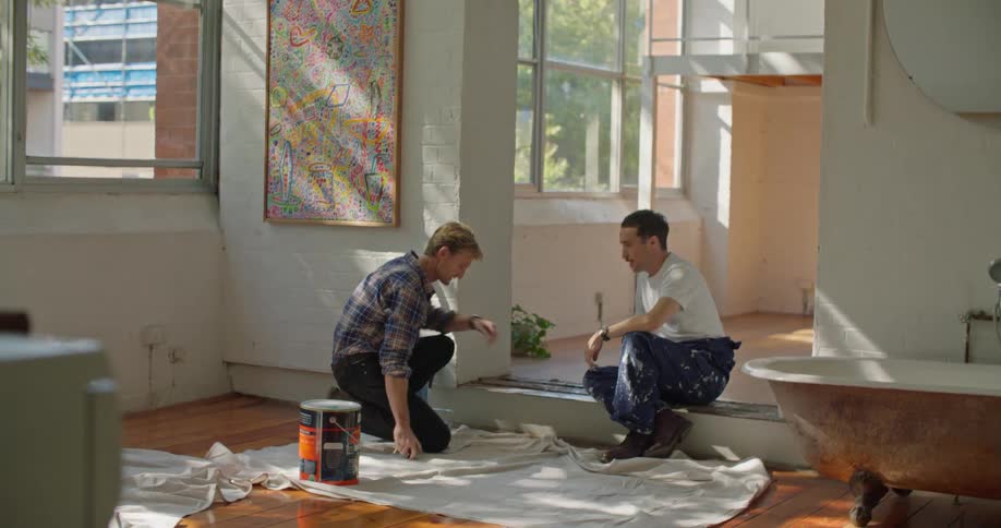 A man sits in a house and talks and points as another man takes measurements and notes