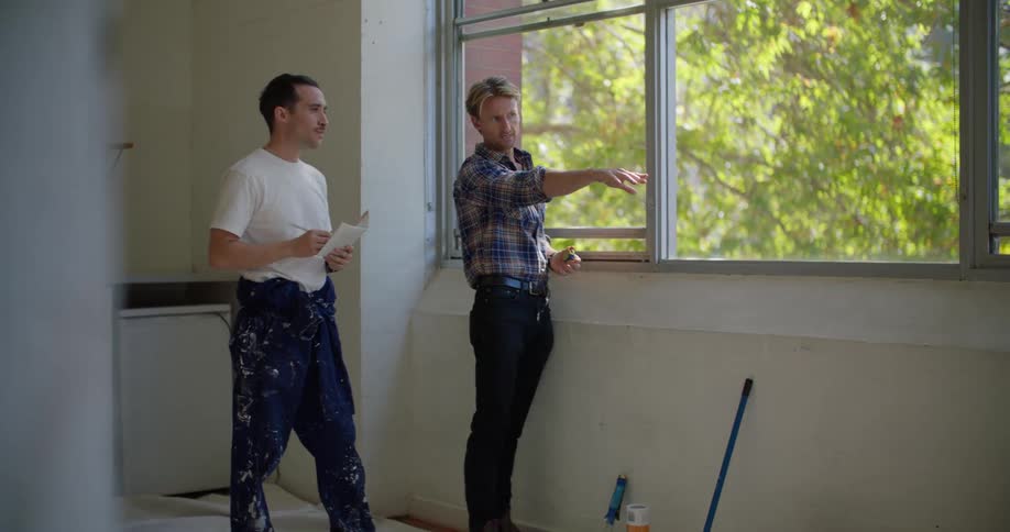 A man gestures across the room while speaking with another man in an unpainted room during the day