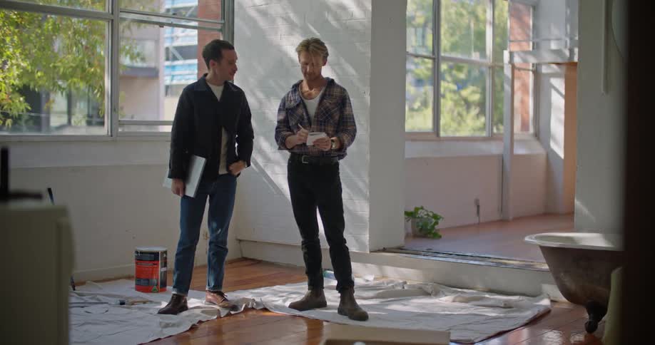 A man writes on a folded sheet of paper while talking with another man standing beside him indoors during the day