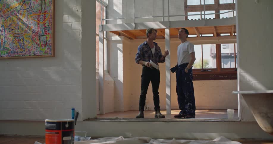 A pair of men gesture at their surroundings as they speak with one another in a building during the day