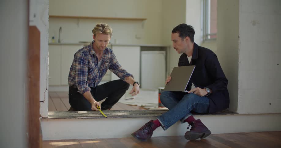 Man sits typing on laptop as another man measures floor area in kitchen