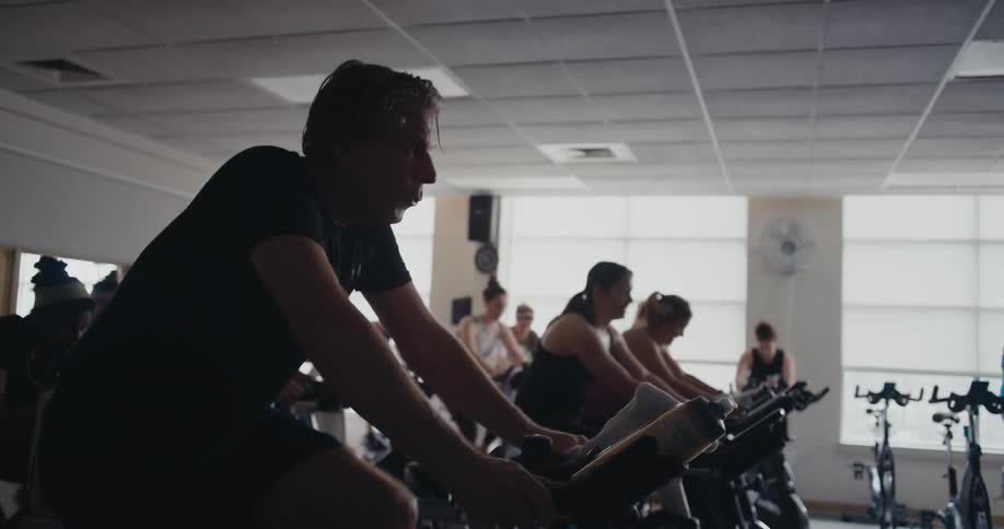 A man exhales as he continues to pedal an exercise bike at the gym in a class full of others