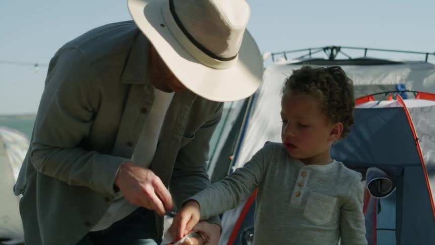 A little boy helps his dad take hotdogs out of a package to place on the campsite grill