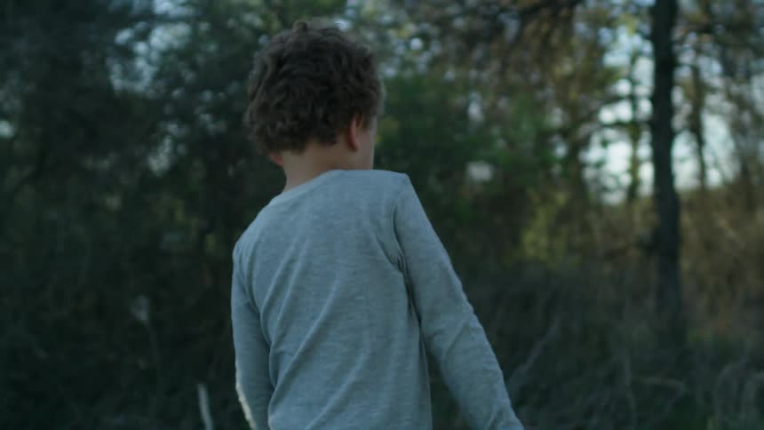 A boy looks down at the ground while walking in the woods