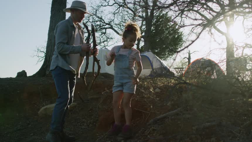 A little girl is helping her father gather firewood near their campsite as the sun shines brightly upon them