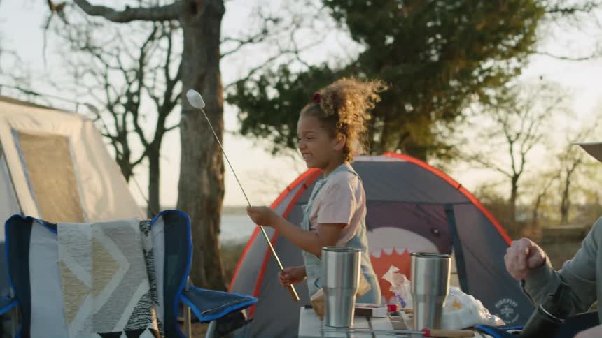 A little girl has a marshmallow on a stick as she walks around the table her parents are at near tents