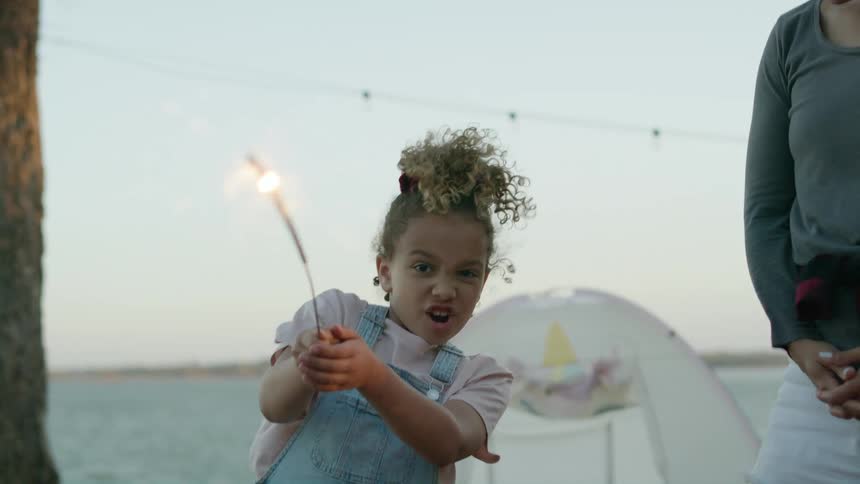 A little girl holds onto a sparkler with both hands as the wind blows off the lake near her tent